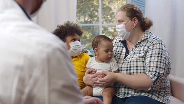Male doctor talking to caucasian mother and mixed-race children at home wearing face mask. Realtime