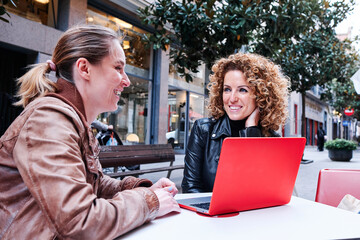 Two women smiling while using laptop sitting on a table outdoors on the street.