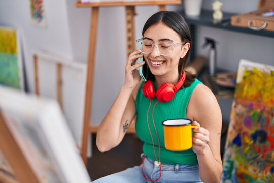 Young hispanic woman artist talking on the smartphone drinking coffee at art studio