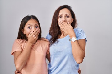 Young mother and daughter standing over white background shocked covering mouth with hands for...