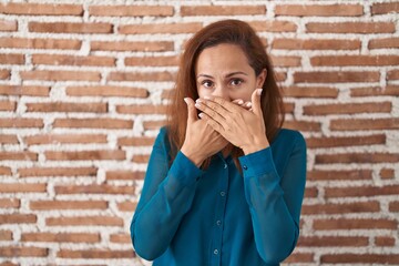 Brunette woman standing over bricks wall shocked covering mouth with hands for mistake. secret...