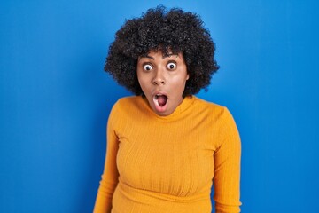 Fototapeta na wymiar Black woman with curly hair standing over blue background in shock face, looking skeptical and sarcastic, surprised with open mouth