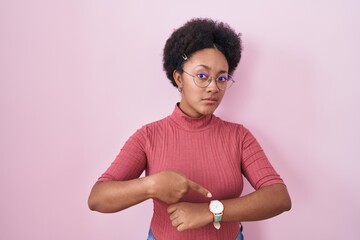 Beautiful african woman with curly hair standing over pink background in hurry pointing to watch...