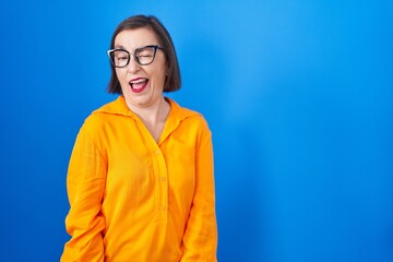 Middle age hispanic woman wearing glasses standing over blue background winking looking at the camera with sexy expression, cheerful and happy face.