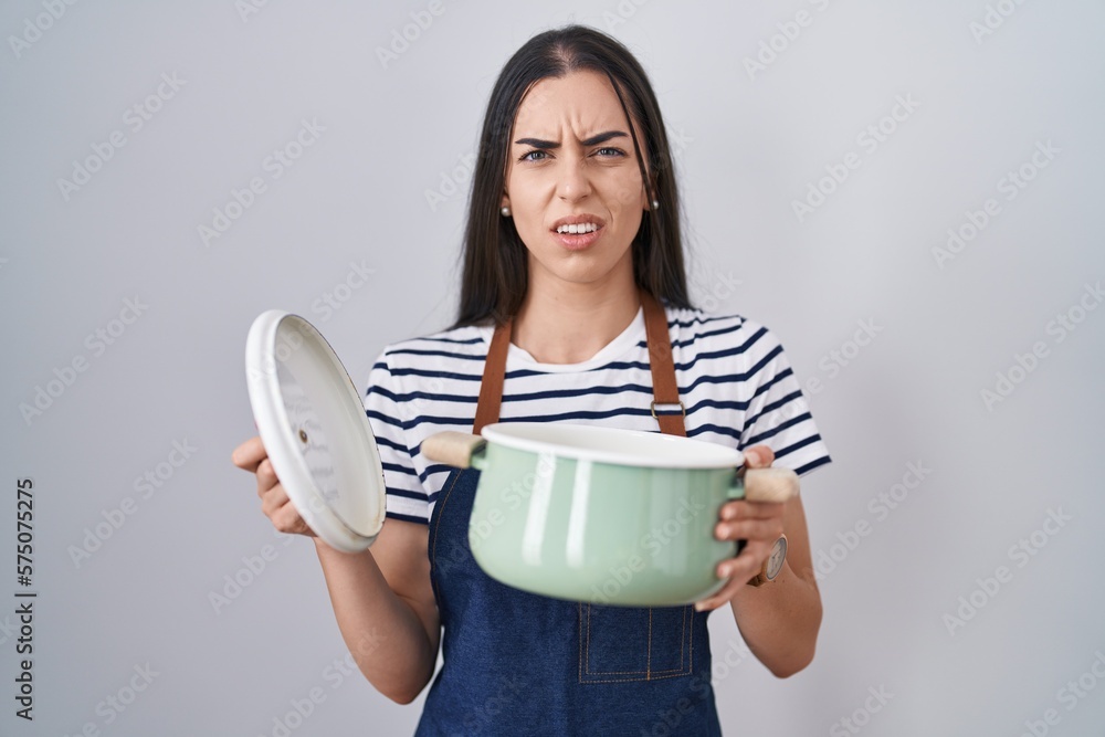 Canvas Prints Young brunette woman wearing apron holding cooking pot clueless and confused expression. doubt concept.