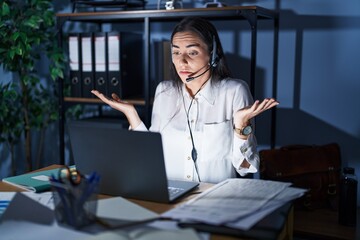 Young brunette woman wearing call center agent headset working late at night clueless and confused...