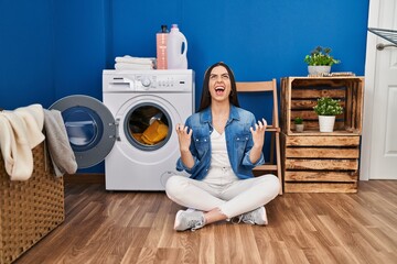 Hispanic woman doing laundry sitting on the floor crazy and mad shouting and yelling with aggressive expression and arms raised. frustration concept.