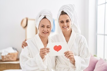Middle age woman and daughter wearing bath robe holding heart card winking looking at the camera...