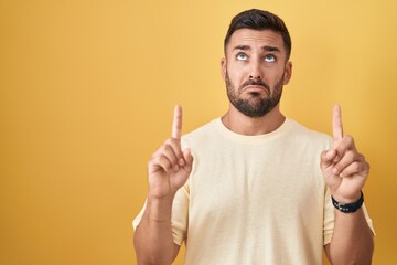 Handsome hispanic man standing over yellow background pointing up looking sad and upset, indicating direction with fingers, unhappy and depressed.