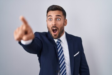 Handsome hispanic man wearing suit and tie pointing with finger surprised ahead, open mouth amazed expression, something on the front