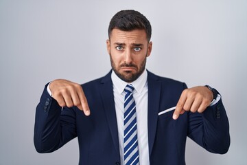 Handsome hispanic man wearing suit and tie pointing down looking sad and upset, indicating direction with fingers, unhappy and depressed.