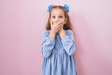 Young little girl standing over pink background shocked covering mouth with hands for mistake....