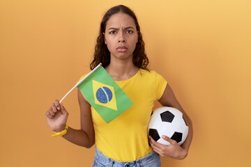 Young hispanic woman holding brazil flag and football ball skeptic and nervous, frowning upset...