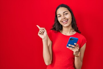 Young brazilian woman using smartphone over red background with a big smile on face, pointing with hand finger to the side looking at the camera.