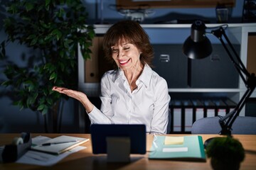 Middle age woman working at the office at night pointing aside with hands open palms showing copy space, presenting advertisement smiling excited happy