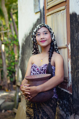 Asian girl dressed in traditional old batik sarongs in front of the wall and holds a clay pot for...