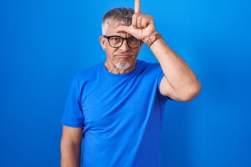 Hispanic man with grey hair standing over blue background making fun of people with fingers on...
