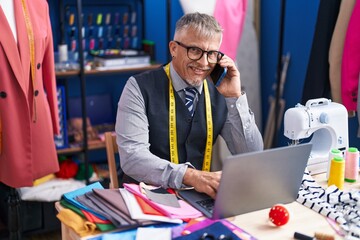 Middle age grey-haired man tailor talking on smartphone using laptop at clothing factory