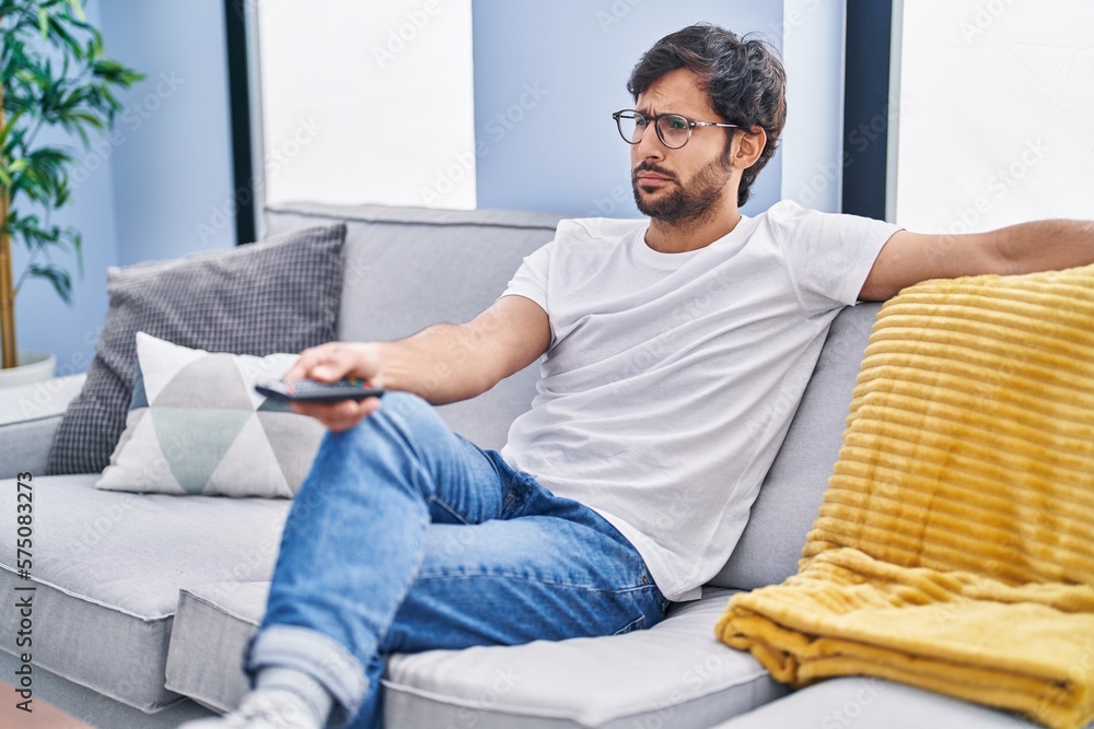 Sticker Handsome latin man holding television remote control depressed and worry for distress, crying angry and afraid. sad expression.