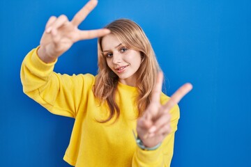 Young caucasian woman standing over blue background smiling with tongue out showing fingers of both hands doing victory sign. number two.