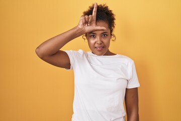 Young hispanic woman with curly hair standing over yellow background making fun of people with fingers on forehead doing loser gesture mocking and insulting.