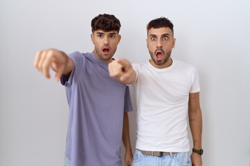 Homosexual gay couple standing over white background pointing with finger surprised ahead, open mouth amazed expression, something on the front