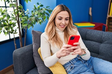 Young blonde woman using smartphone sitting on sofa at home