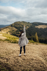 Happy girl stands in the mountains, enjoys the beautiful view and raises her hand up