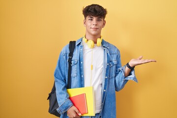 Hispanic teenager wearing student backpack and holding books smiling cheerful presenting and pointing with palm of hand looking at the camera.