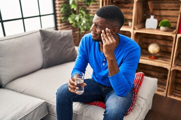 Young african american man with headache holding glass of water at home