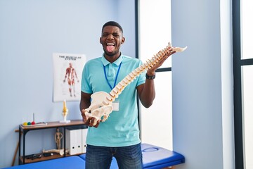 African american man holding anatomical model of spinal column sticking tongue out happy with funny...