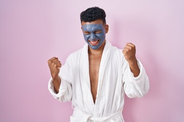 Young hispanic man wearing beauty face mask and bath robe very happy and excited doing winner...