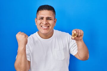 Fototapeta premium Hispanic young man holding virtual currency bitcoin screaming proud, celebrating victory and success very excited with raised arms