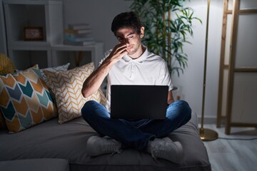 Young hispanic man using laptop at home at night smelling something stinky and disgusting,...