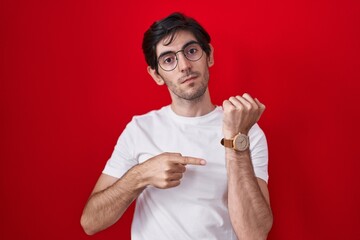 Young hispanic man standing over red background in hurry pointing to watch time, impatience,...