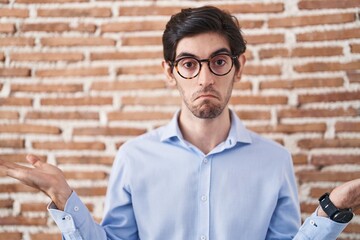 Young hispanic man standing over brick wall background clueless and confused expression with arms...