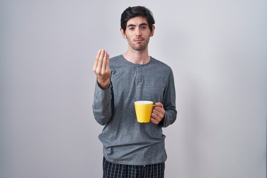 Young hispanic man wearing pajama drinking a cup of coffee doing italian gesture with hand and fingers confident expression
