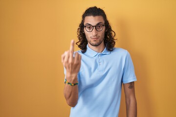 Young hispanic man standing over yellow background showing middle finger, impolite and rude fuck...