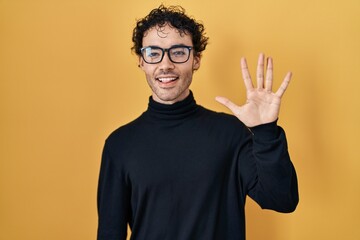 Hispanic man standing over yellow background showing and pointing up with fingers number five while smiling confident and happy.