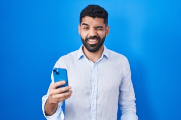 Hispanic man with beard using smartphone typing message winking looking at the camera with sexy expression, cheerful and happy face.