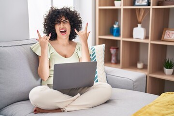 Young brunette woman with curly hair using laptop sitting on the sofa at home shouting with crazy expression doing rock symbol with hands up. music star. heavy concept.