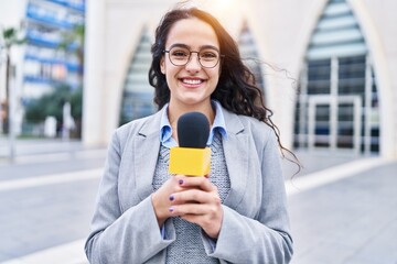 Young brunette woman holding reporter microphone looking positive and happy standing and smiling...