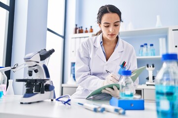 Young hispanic woman wearing scientist uniform measuring liquid at laboratory