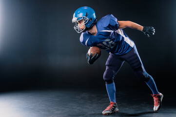 Fototapeta na wymiar Full length portrait of a man in a blue american football uniform against a black background. Sportsman in a helmet with a ball. 