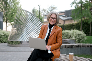 pretty gray-haired business woman of mature age works with a laptop on a bench in the park
