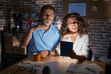 Fototapeta premium Middle age hispanic couple using touchpad sitting on the table at night cutting throat with hand as knife, threaten aggression with furious violence