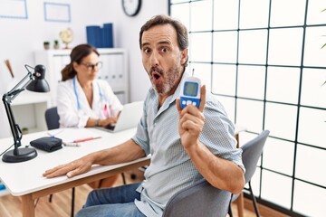Middle age man at doctor clinic holding glucose meter device scared and amazed with open mouth for...