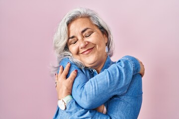 Middle age woman with grey hair standing over pink background hugging oneself happy and positive,...