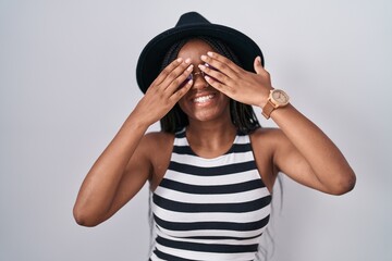 Young african american with braids wearing hat and sunglasses covering eyes with hands smiling cheerful and funny. blind concept.