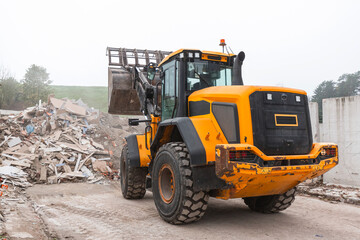 Yellow wheel loader, with lifted scrap grapple, moving along the recycling center area in process handling dumped waste